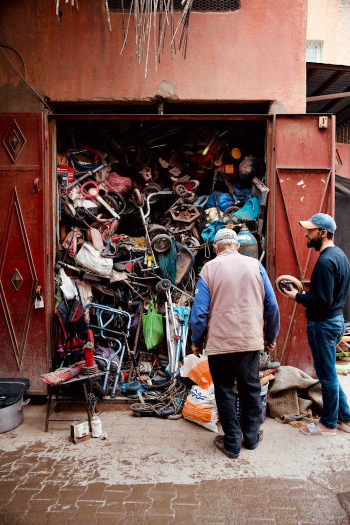men-standing-in-front-of-a-storage-full-of-trash-15193736 Two men inspecting a workshop with a cluttered collection of discarded items and tools.